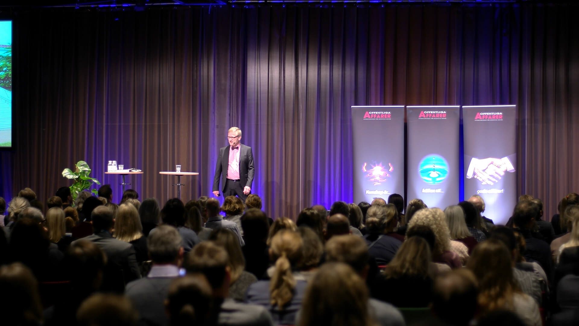 Niklas on stage in a pink shirt and black blazer, speaking to a full room